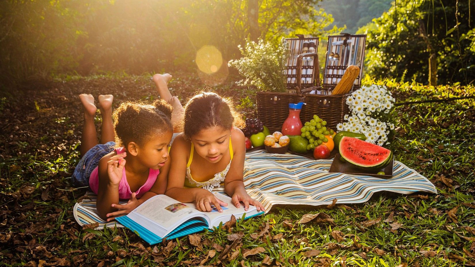 Two kids laying down to read a book on a blanket on the grass, while on a picnic